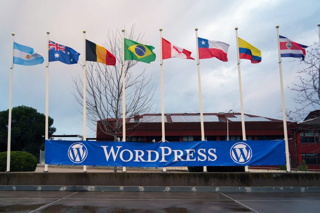 A row of nine flags representing different countries, including Argentina, Australia, Belgium, Brazil, Canada, Chile, Columbia, Costa Rica, are displayed on tall poles against a backdrop of a building with solar panels on the roof. A large blue banner with the word “WordPress” is positioned below the flags.
