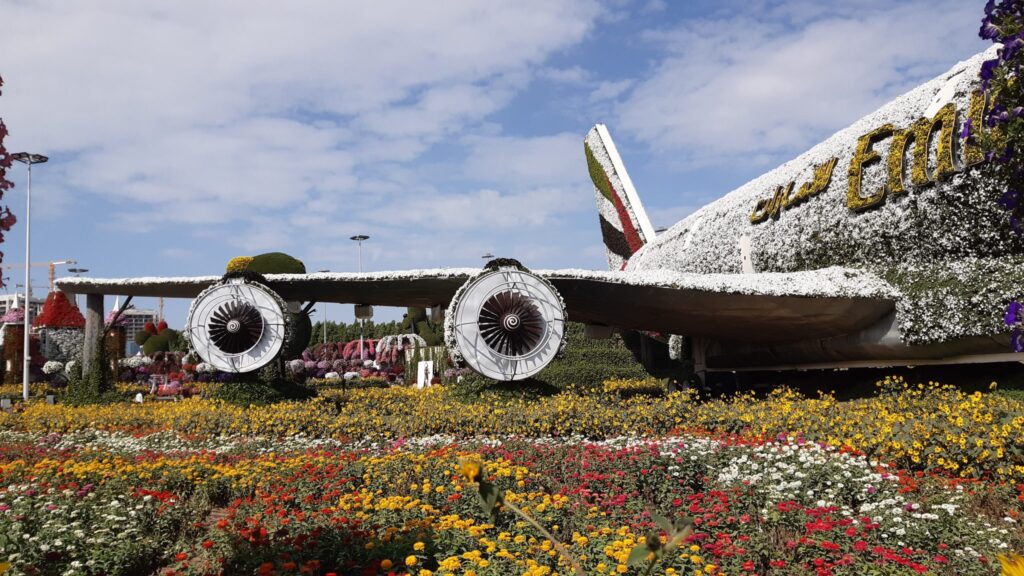 A large airplane structure covered in white and yellow flowers with the letters “Emi” visible on the side, set amidst a vibrant garden with colorful flowers under a partly cloudy sky.