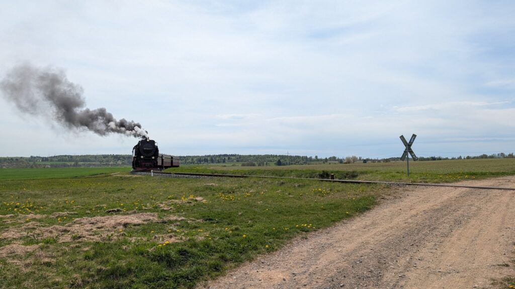 The Brockenbahn steam train travels through a wide green landscape, leaving a trail of dark smoke as it approaches a rural railway crossing.