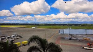 A view of an airport runway under a blue sky filled with fluffy clouds. In the foreground, there is a boarding gate with a staircase leading down, and several ground service vehicles including luggage carts and tugs.